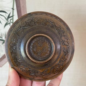 May include: A bronze-colored, antique bowl with intricate floral and leaf carvings around the rim. The center features a circular design with Chinese characters. The bowl is held in a person's hand.