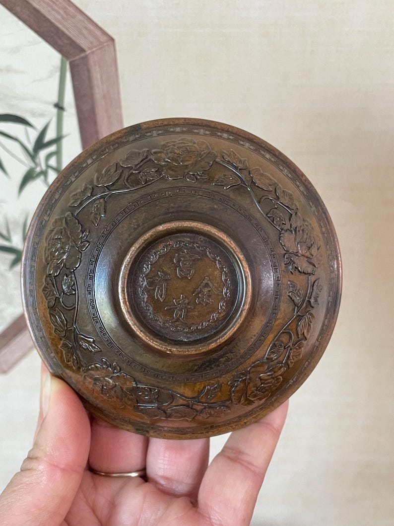May include: A bronze-colored, antique bowl with intricate floral and leaf carvings around the rim. The center features a circular design with Chinese characters. The bowl is held in a person's hand.
