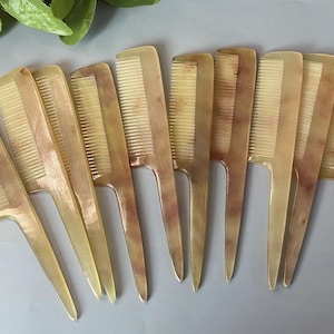 Puede incluir: Una colección de diez peines de cuerno, cada uno con mango y dientes finos. Los peines son de color marrón claro con veteado marrón oscuro, dispuestos sobre una superficie gris claro. Los peines son para el cuidado del cabello.