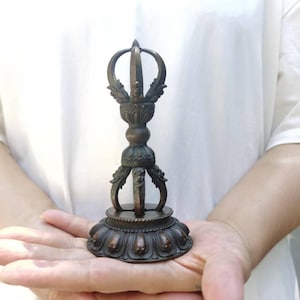 Pode incluir: Vajra de cor bronze, um objeto ritual, segurado na palma da mão aberta. O Vajra apresenta um design detalhado com uma base redonda, uma coluna central e pontas curvas e ornamentadas no topo. O objeto tem cerca de 15 cm de altura.