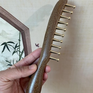 Puede incluir: Un peine de madera con un mango curvo y púas de metal dorado. El peine está hecho de madera marrón oscuro y tiene un acabado liso y pulido. Las púas están espaciadas uniformemente y tienen puntas redondeadas. El peine es para el cuidado del cabello.
