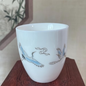 May include: White ceramic teacup with a rounded shape. The cup features a blue and gold illustration of two cranes in flight. The design includes stylized clouds and flowing ribbons. The cup is displayed on a dark wooden surface.