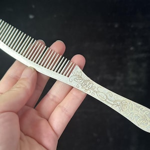 May include: Silver comb with a curved handle, decorated with floral engravings. The handle features roses and leaves. The comb's teeth are evenly spaced. The comb is held against a dark background.