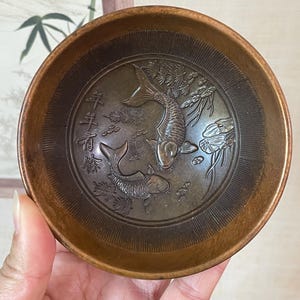 May include: A bronze-colored, round bowl with an embossed design of two koi fish and aquatic plants. The bowl's interior features intricate details, and the rim has a smooth, polished finish. The bowl is held in a hand, showcasing its size and shape.