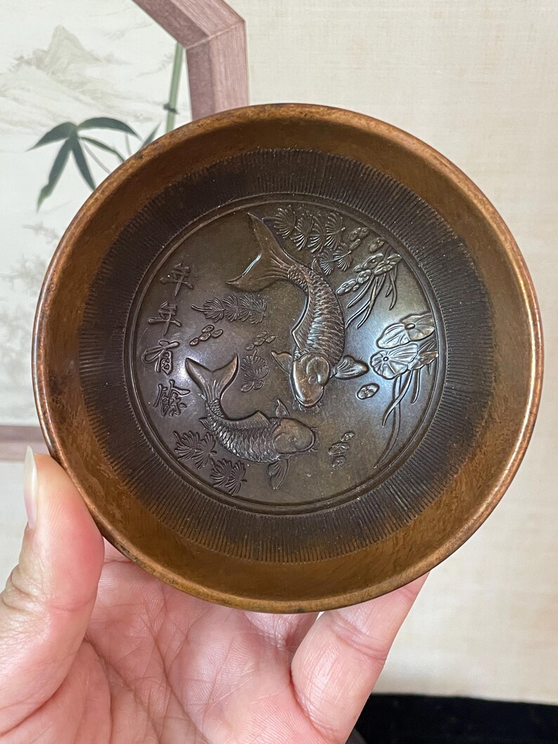May include: A bronze-colored, round bowl with an embossed design of two koi fish and aquatic plants. The bowl's interior features intricate details, and the rim has a smooth, polished finish. The bowl is held in a hand, showcasing its size and shape.
