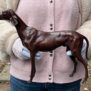 Puede incluir: Una escultura de perro de color bronce, de pie con un cuerpo delgado y alargado y orejas puntiagudas. La escultura está sostenida por una persona que lleva un suéter rosa y un abrigo color crema. La forma del perro es detallada, mostrando sus costillas y su físico delgado.