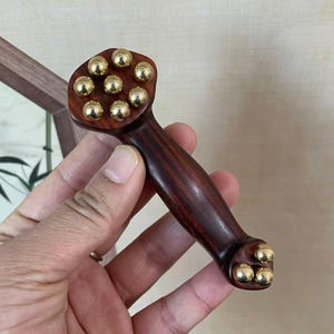 Op de afbeelding: Een donkerroodbruin houten massagegereedschap met goudkleurige metalen bollen. Het gereedschap heeft een afgeronde kop met negen bollen en een handvat met drie bollen aan het uiteinde. De houtnerf is zichtbaar.