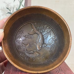 May include: A small, round, bronze-colored bowl with an embossed design of two koi fish and Chinese characters. The bowl has a slightly raised rim and a detailed interior. The fish are surrounded by aquatic plants.