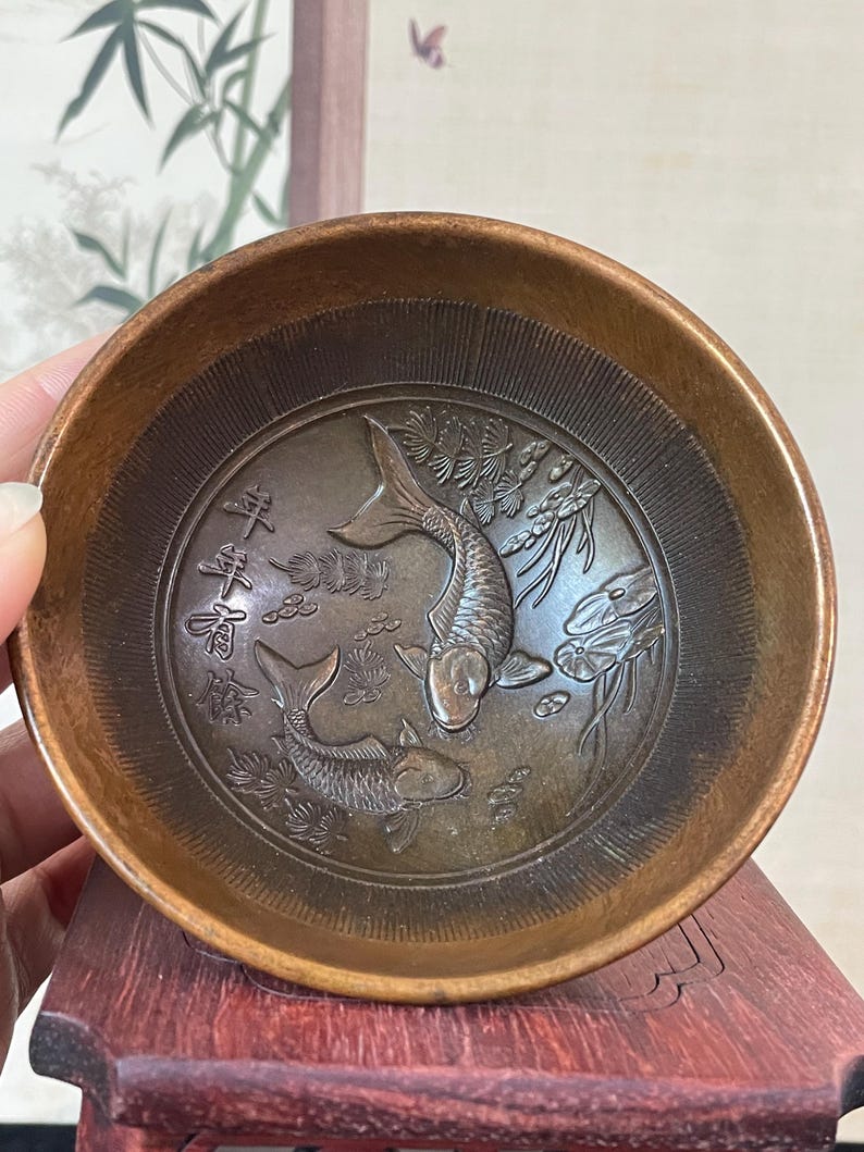 May include: A small, round, bronze-colored bowl with an embossed design of two koi fish and Chinese characters. The bowl has a slightly raised rim and a detailed interior. The fish are surrounded by aquatic plants.