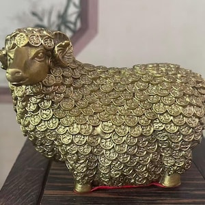 May include: A gold-coloured ram sculpture with a textured surface, possibly made of brass or bronze. The ram's body is covered in coin-like details, and it has curved horns and a detailed face. The sculpture is a decorative item.
