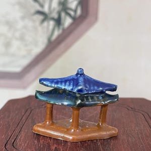 May include: A miniature ceramic pagoda with a two-tiered roof in shades of blue and brown. The pagoda has a square base and is supported by four brown pillars. The background is blurred, with a hint of a painting.