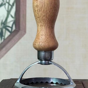 Peut inclure: Un ustensile de cuisine avec un manche en bois et un bord dentelé en acier inoxydable. Le manche est brun clair et les parties métalliques sont argentées. L'outil est probablement destiné à couper ou presser des aliments.