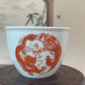 Op de afbeelding: Een witte keramische theekop met een rood draak- en feniksontwerp. Het kunstwerk is gecentreerd op de kop, met de draak en feniks tegenover elkaar. De kop staat op een donkerhouten standaard.