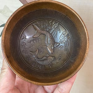 May include: A bronze-colored bowl with an embossed design featuring two koi fish and Chinese characters. The bowl has a circular shape with a slightly flared rim and a detailed interior.