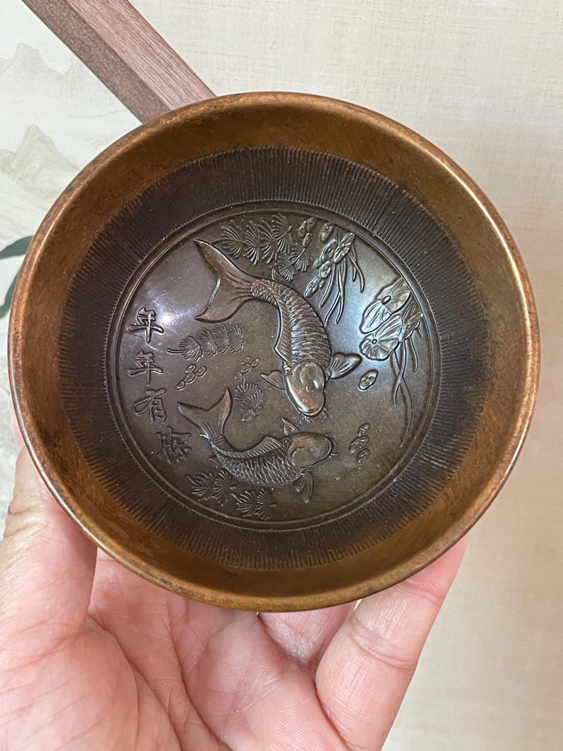 May include: A bronze-colored bowl with an embossed design featuring two koi fish and Chinese characters. The bowl has a circular shape with a slightly flared rim and a detailed interior.