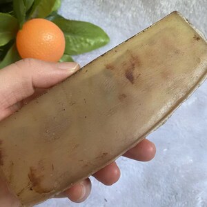May include: A rectangular, light brown soap bar with a slightly translucent appearance. The soap has dark brown spots and is held in a hand. An orange fruit and green leaves are in the background.
