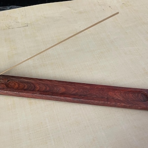 May include: A rectangular, dark reddish-brown wooden incense holder with a long, narrow trough and a small, angled block at one end to hold the incense stick. A single, thin, tan incense stick is in place. The holder is approximately 12 inches long.