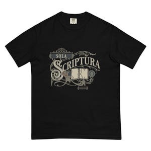 Op de afbeelding: Zwart T-shirt met een decoratief ontwerp met de woorden "SOLA SCRIPTURA" in een sierlijk lettertype. Het ontwerp bevat een open boek en krullen in grijstinten en beige.