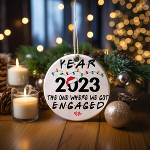 May include: White ceramic Christmas ornament with the text "Year 2023 The One Where We Got Engaged" and a small Santa hat on the 2. The ornament is surrounded by pine cones and candles.