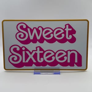 Op de afbeelding: Een rechthoekig bord met een gouden rand en de woorden "Sweet Sixteen" in roze, gestileerd lettertype. Het bord wordt weergegeven op een transparante plastic standaard tegen een witte achtergrond. Ideaal voor een verjaardagsfeest.