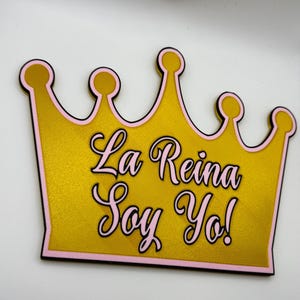 Op de afbeelding: Een goudkleurige kroon met een roze rand en de tekst "La Reina Soy Yo!" in roze script. De kroon heeft een zwarte omtrek en staat tegen een witte achtergrond.