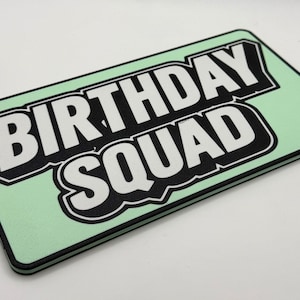 Op de afbeelding: Een rechthoekig, mintgroen item met de tekst "BIRTHDAY SQUAD" in vette witte letters met een zwarte omlijning. Het heeft een zwarte rand, mogelijk een decoratief bord.