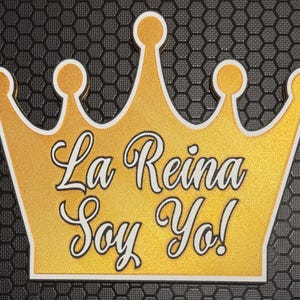 Op de afbeelding: Een goudkleurige kroon met de tekst "La Reina Soy Yo!" in zwart script. De kroon heeft een witte rand en een getextureerd gouden oppervlak. De achtergrond is een donkergrijs, zeshoekig patroon.