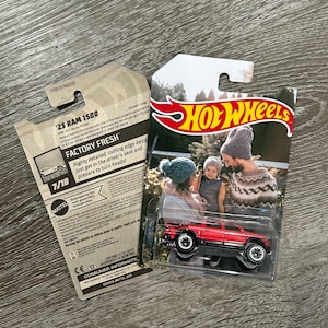 May include: A red 2023 Ram 1500 pickup truck toy car with black wheels and a black interior. The toy car is on a white background with a family in the background. The packaging has the words "Hot Wheels" and "Factory Fresh" on it.