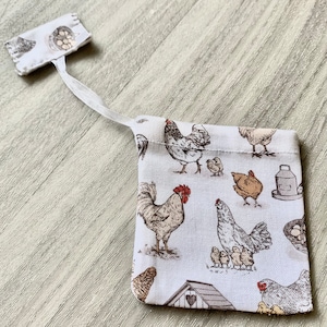 Puede incluir: Una bolsa de tela blanca con cordón de ajuste, con estampado de pollos y huevos. La bolsa tiene un cordón de ajuste blanco y una pequeña etiqueta de tela blanca con estampado de pollos y huevos.