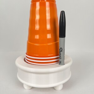 May include: White plastic cup holder with three legs, holding a stack of orange plastic cups and a black Sharpie fine point marker.