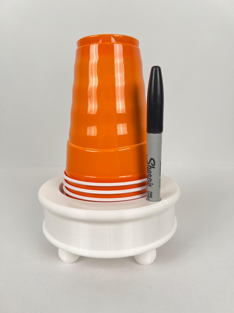 May include: White plastic cup holder with three legs, holding a stack of orange plastic cups and a black Sharpie fine point marker.