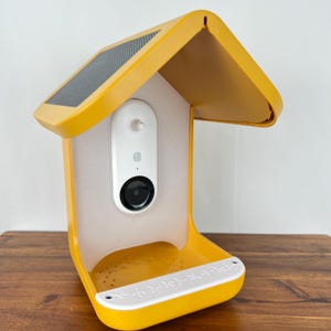 May include: A yellow birdhouse with a solar panel on the roof and a white security camera mounted inside. The birdhouse has a white plastic base with the words "Bird Buddy" printed on it.