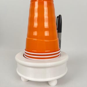 May include: White plastic cup holder with three legs and a round top. An orange plastic cup is sitting on top of the holder.