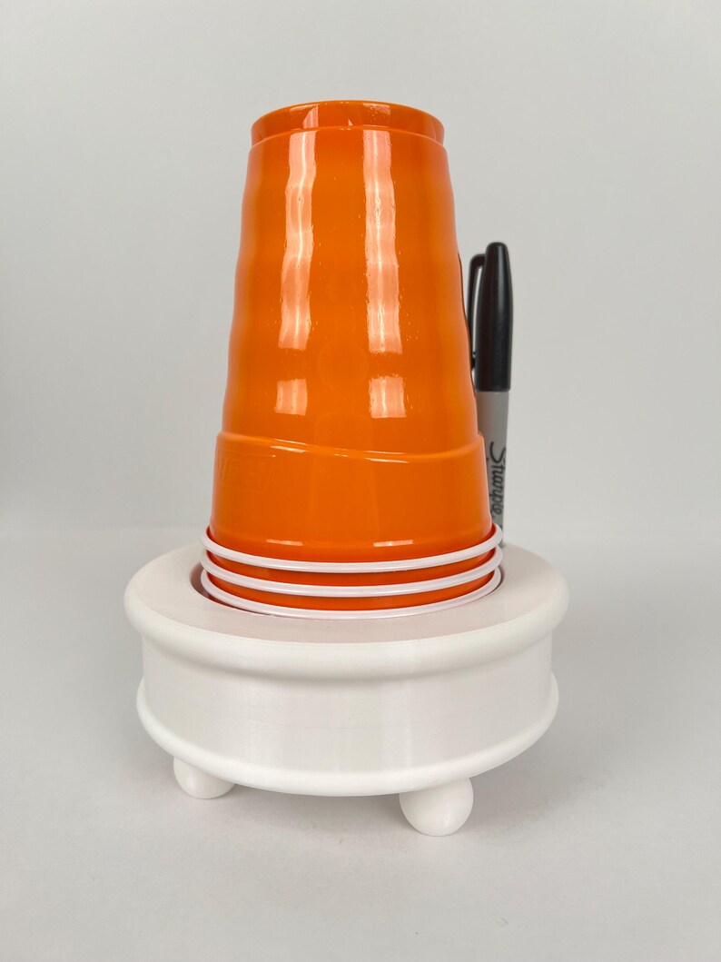 May include: White plastic cup holder with three legs and a round top. An orange plastic cup is sitting on top of the holder.