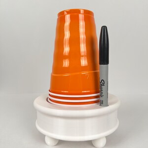 May include: White plastic cup holder with three legs and a black Sharpie marker in the holder. The holder has a round top and a cup is sitting on top of it.