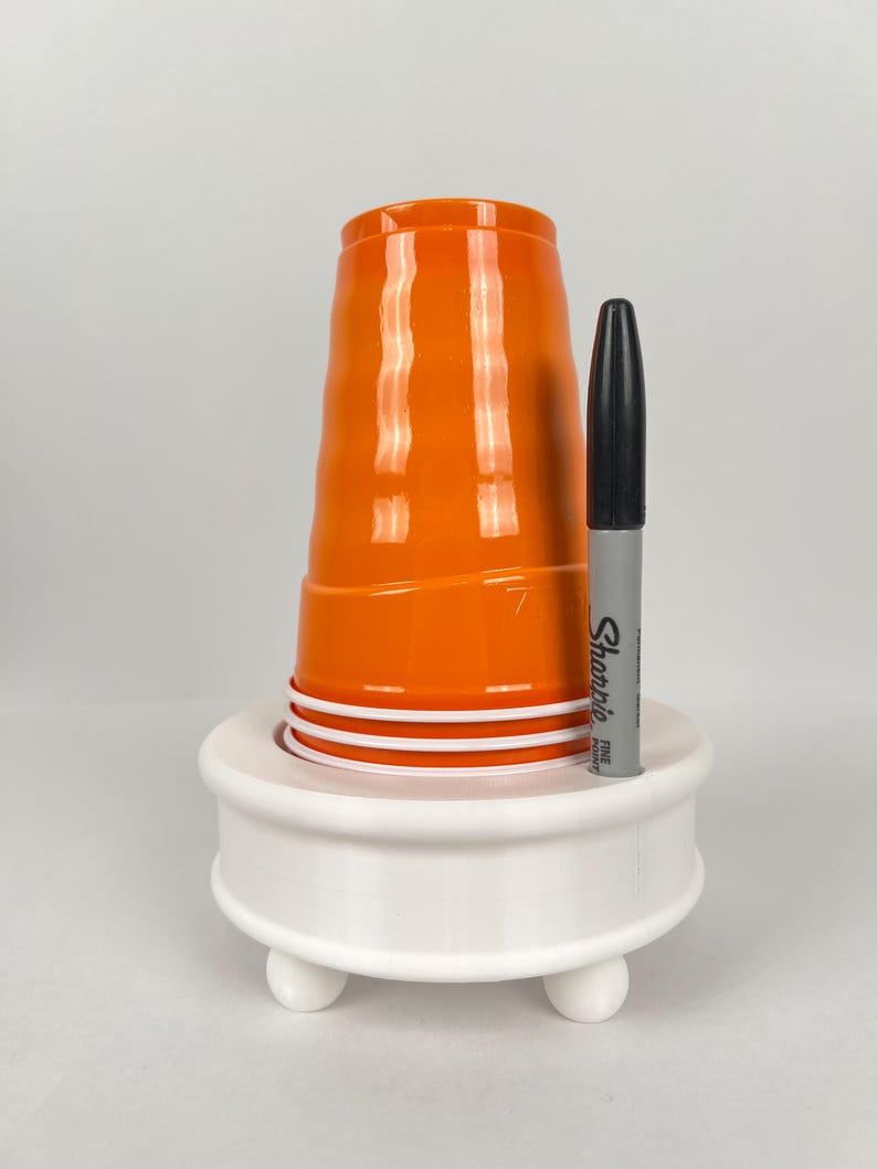 May include: White plastic cup holder with three legs and a black Sharpie marker in the holder. The holder has a round top and a cup is sitting on top of it.