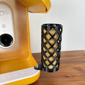 May include: A black, lattice-patterned cylinder holds a light brown, cylindrical object. The cylinder is attached to a yellow and white device with a camera and a small dish. The device is on a wooden surface.