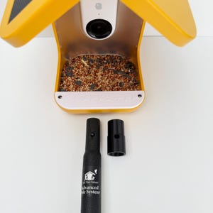 May include: A yellow bird feeder with a built-in camera and a white base, filled with birdseed. The feeder has a black pole and a black connector. The text "Advanced Pole System" is visible on the pole.