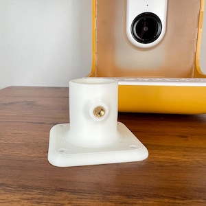 May include: A white plastic camera mount with a square base and a cylindrical top. A small gold-colored screw is visible on the side. The background includes a yellow and white camera housing and a wooden surface.