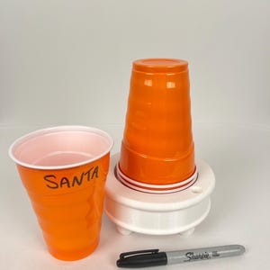 May include: Two orange plastic cups, one with the word "SANTA" written on it, are stacked on a white plastic cup holder. A black Sharpie marker is lying on a white surface.