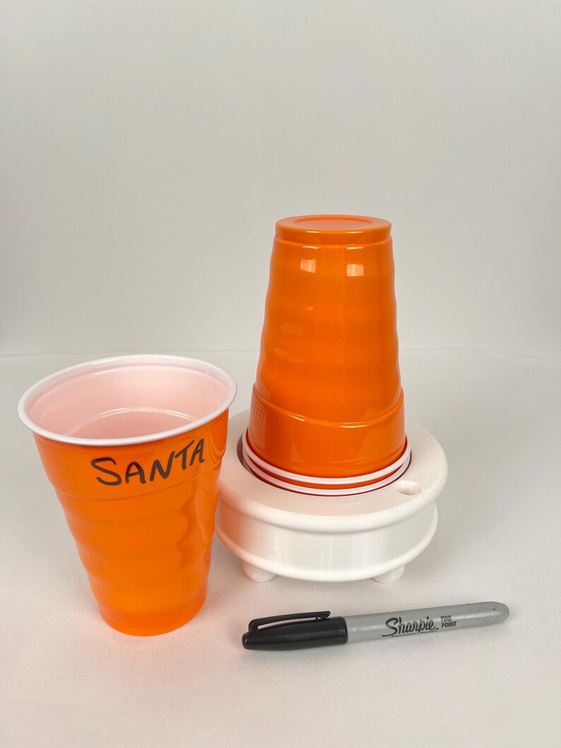 May include: Two orange plastic cups, one with the word "SANTA" written on it, are stacked on a white plastic cup holder. A black Sharpie marker is lying on a white surface.