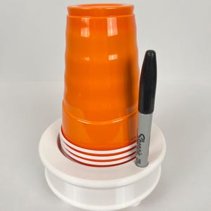 May include: A white plastic cup holder with a stack of orange plastic cups and a black Sharpie marker.