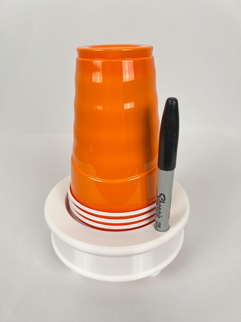 May include: A white plastic cup holder with a stack of orange plastic cups and a black Sharpie marker.