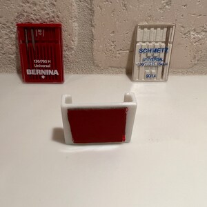 May include: Two packs of sewing machine needles, one red and one clear, with the brand names "BERNINA" and "SCHMETZ" printed on them. The packs are sitting on a white surface next to a white plastic needle holder with a red adhesive pad.