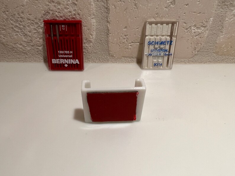 May include: Two packs of sewing machine needles, one red and one clear, with the brand names "BERNINA" and "SCHMETZ" printed on them. The packs are sitting on a white surface next to a white plastic needle holder with a red adhesive pad.