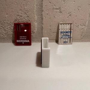 May include: Three packages of sewing machine needles. The package on the left is red and says "130/705 H Universal BERNINA". The package on the right is clear and says "SCHMETZ UNIVERSAL 130/705 H 135x5  90114". The package in the middle is white.