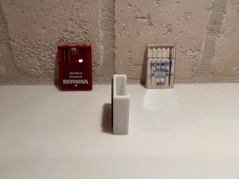 May include: Three packages of sewing machine needles. The package on the left is red and says "130/705 H Universal BERNINA". The package on the right is clear and says "SCHMETZ UNIVERSAL 130/705 H 135x5  90114". The package in the middle is white.