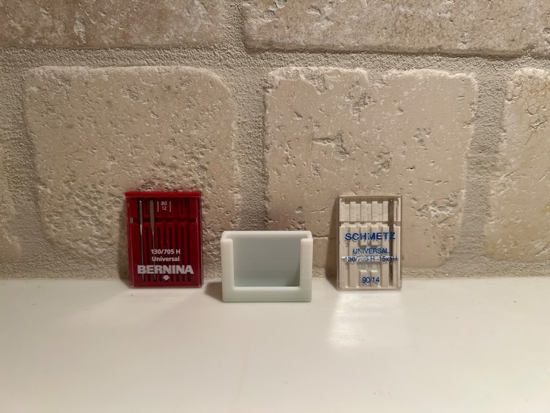 May include: Two packs of sewing machine needles. The pack on the left is red and says "BERNINA TRUE-LOVER 130/705 H Universal". The pack on the right is clear and says "SCHMETZ UNIVERSAL 130/705 H-16x1+ 9014".