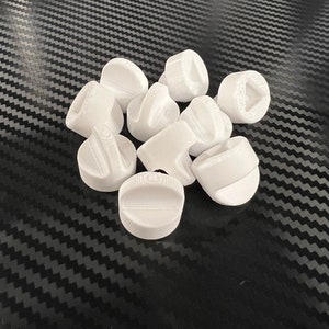 May include: A group of ten white plastic knobs with different designs. The knobs are arranged in a pile on a black and white patterned surface.