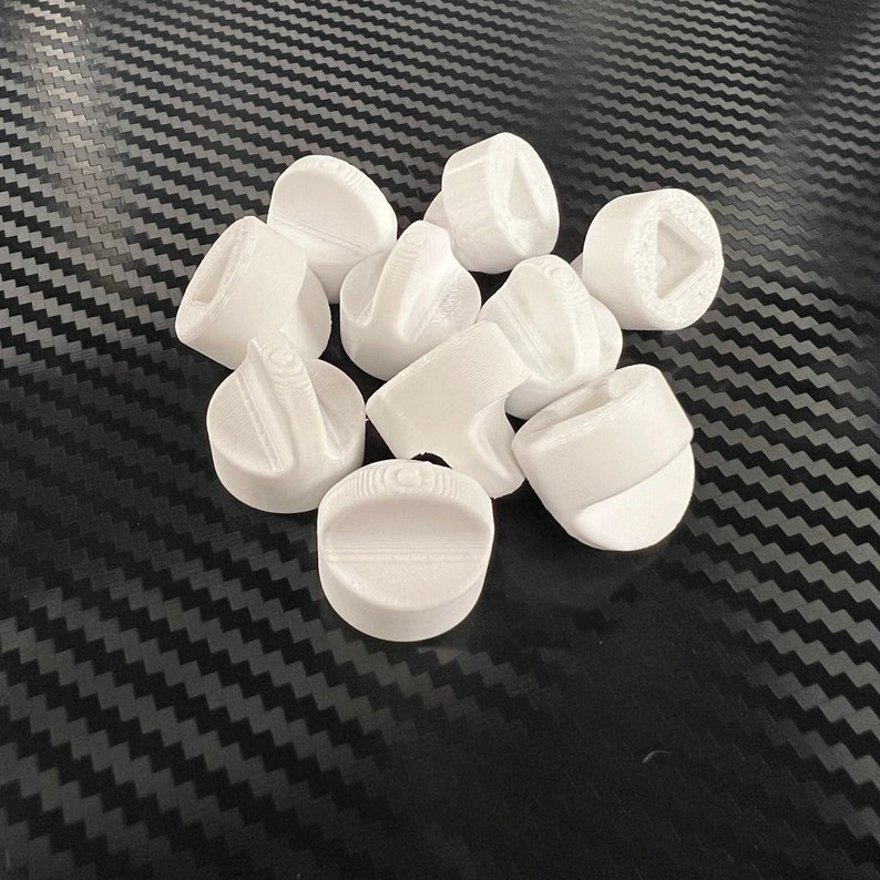 May include: A group of ten white plastic knobs with different designs. The knobs are arranged in a pile on a black and white patterned surface.